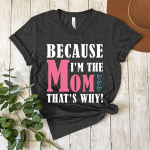 May include: A dark gray t-shirt with the text "Because I'm the Mom That's Why!" in white. The word "Mom" is in pink with small blue hearts.