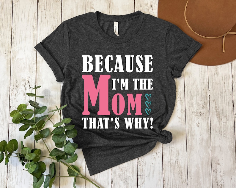 May include: A dark gray t-shirt with the text "Because I'm the Mom That's Why!" in white. The word "Mom" is in pink with small blue hearts.