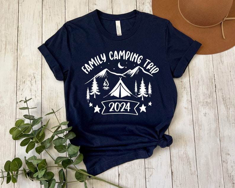 Pu&ograve; includere: Una maglietta blu navy con un design grafico bianco di una scena di campeggio. Il design include una tenda, montagne, alberi, un fal&ograve; e una luna crescente. Il testo "Family Camping Trip 2024" &egrave; incluso nel design.