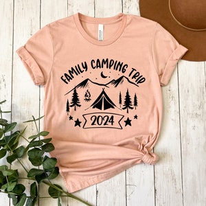 Pu&ograve; includere: Una maglietta color pesca con un design grafico nero di montagne, una tenda, un fal&ograve;, stelle e il testo "Family Camping Trip 2024".