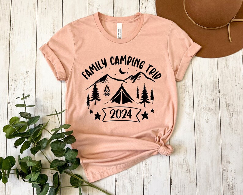 Pu&ograve; includere: Una maglietta color pesca con un design grafico nero di montagne, una tenda, un fal&ograve;, stelle e il testo "Family Camping Trip 2024".
