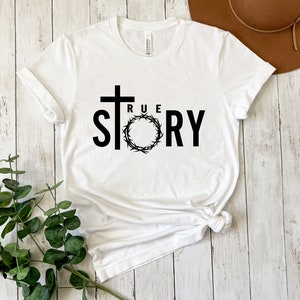 Op de afbeelding: Wit T-shirt met een zwart kruis en de woorden "TRUE STORY" gedrukt op de voorkant. Het kruis is omringd door een doornenkroon.