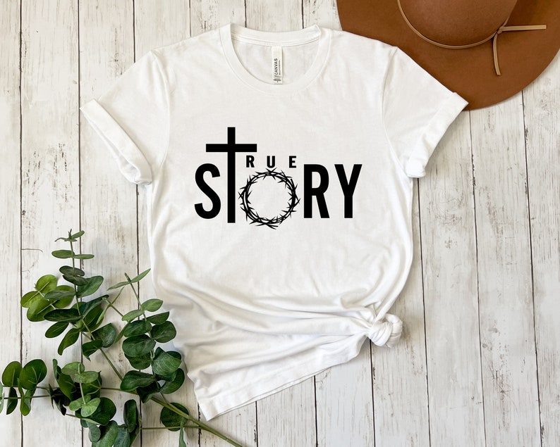 Op de afbeelding: Wit T-shirt met een zwart kruis en de woorden "TRUE STORY" gedrukt op de voorkant. Het kruis is omringd door een doornenkroon.