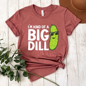 Peut inclure: Un t-shirt marron avec le texte "I'm kind of a big dill" en blanc. Un cornichon de dessin anim&eacute; avec des lunettes de soleil est &agrave; droite du texte.
