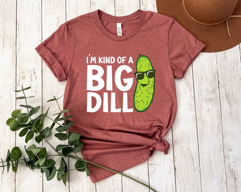 Peut inclure: Un t-shirt marron avec le texte "I'm kind of a big dill" en blanc. Un cornichon de dessin anim&eacute; avec des lunettes de soleil est &agrave; droite du texte.