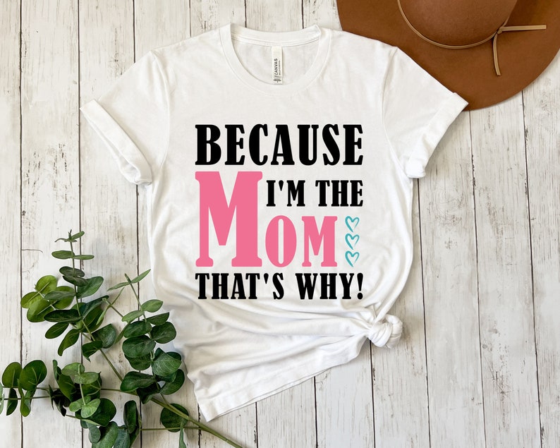 May include: White t-shirt with black text that reads "Because I'm the Mom That's Why!" The word "Mom" is in pink.