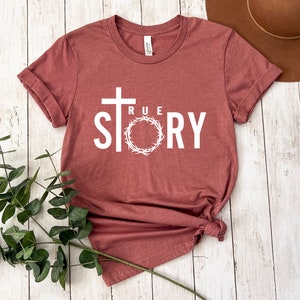 Op de afbeelding: Een maronkleurig T-shirt met een wit kruis, een doornenkroon en de tekst "TRUE STORY".