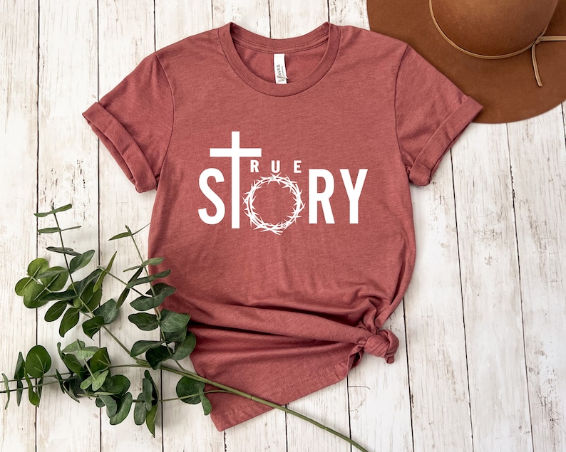 Op de afbeelding: Een maronkleurig T-shirt met een wit kruis, een doornenkroon en de tekst "TRUE STORY".
