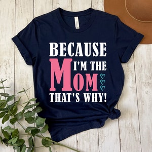 May include: Navy blue t-shirt with white text that reads "Because I'm the Mom That's Why!" The word "Mom" is in pink with teal hearts.