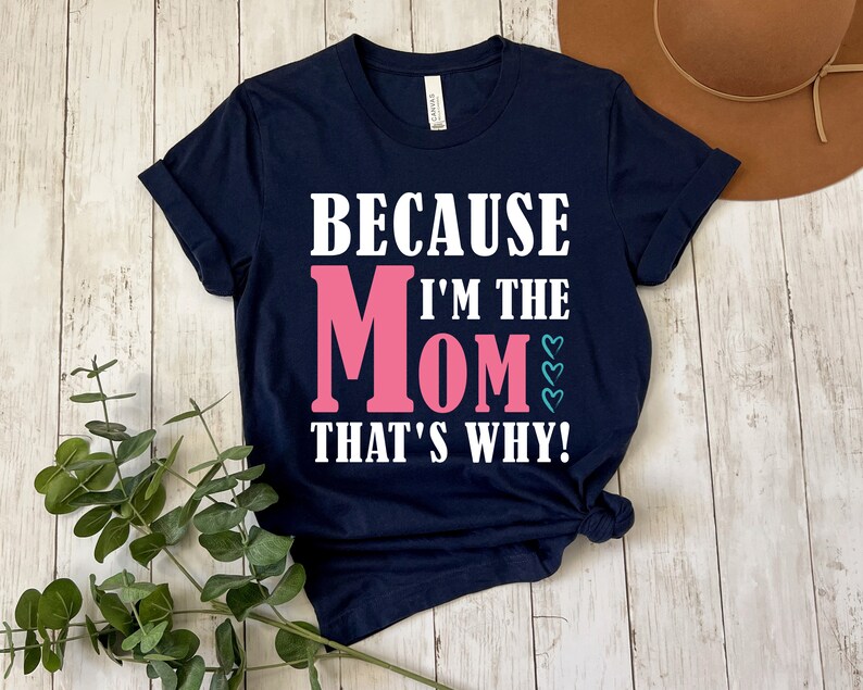 May include: Navy blue t-shirt with white text that reads "Because I'm the Mom That's Why!" The word "Mom" is in pink with teal hearts.