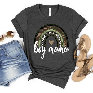 K&ouml;nnte beinhalten: Ein dunkelgraues T-Shirt mit einem Regenbogen-Design aus Tarnmuster, Leopardenmuster und Punkten. Der Regenbogen hat ein kleines Herz in der Mitte und den Text "boy mama" darunter.