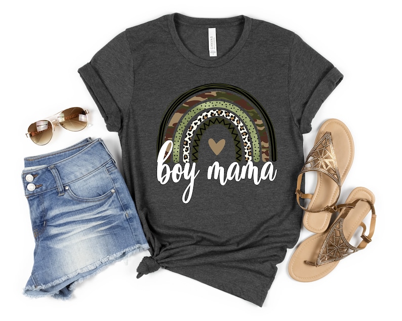 K&ouml;nnte beinhalten: Ein dunkelgraues T-Shirt mit einem Regenbogen-Design aus Tarnmuster, Leopardenmuster und Punkten. Der Regenbogen hat ein kleines Herz in der Mitte und den Text "boy mama" darunter.