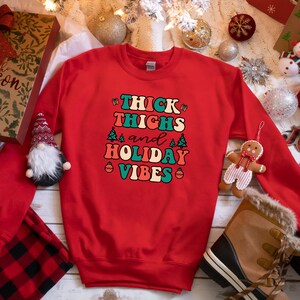 May include: Red Christmas sweatshirt with the text "Thick Thighs and Holiday Vibes" in a retro font with green, blue, and orange accents. The text is surrounded by small Christmas trees and ornaments.