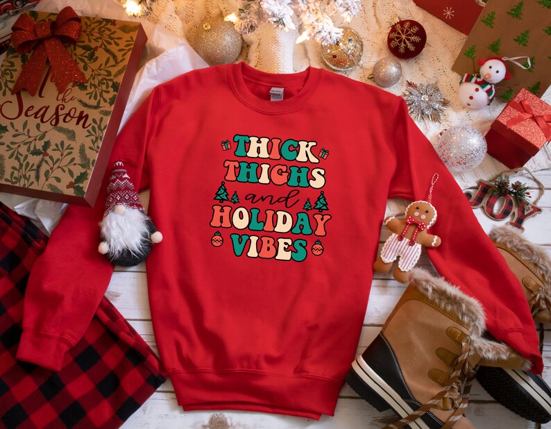 May include: Red Christmas sweatshirt with the text "Thick Thighs and Holiday Vibes" in a retro font with green, blue, and orange accents. The text is surrounded by small Christmas trees and ornaments.