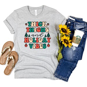 May include: A gray t-shirt with the text "THICK THIGHS and HOLIDAY VIBES" in colorful retro lettering. The shirt is styled with blue jeans, a coffee cup, and sunflowers.