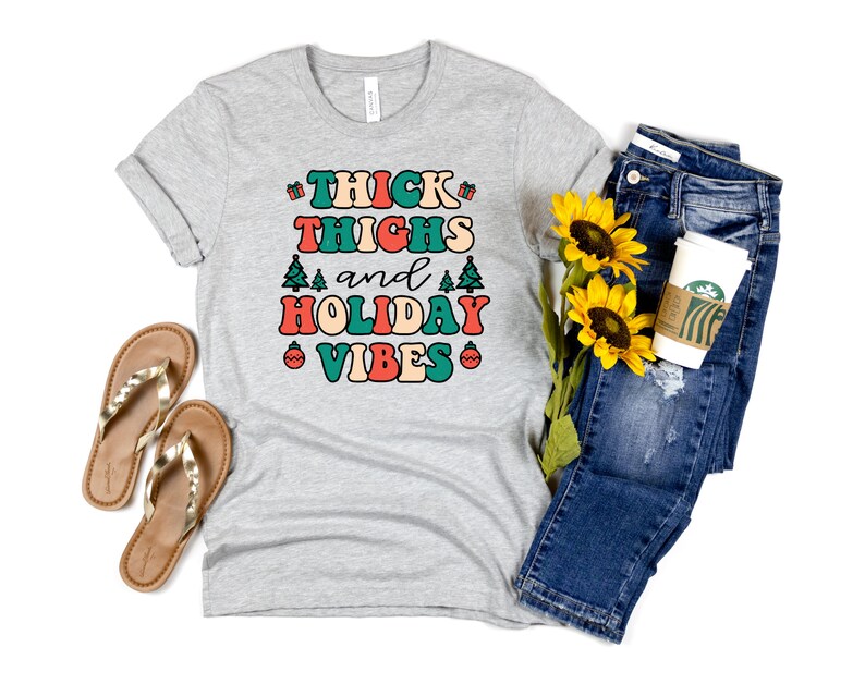 May include: A gray t-shirt with the text "THICK THIGHS and HOLIDAY VIBES" in colorful retro lettering. The shirt is styled with blue jeans, a coffee cup, and sunflowers.