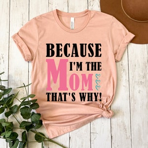 May include: A peach colored t-shirt with the text "Because I'm the Mom That's Why!" in black and pink lettering. The word "Mom" is in pink and has three small pink hearts above it.