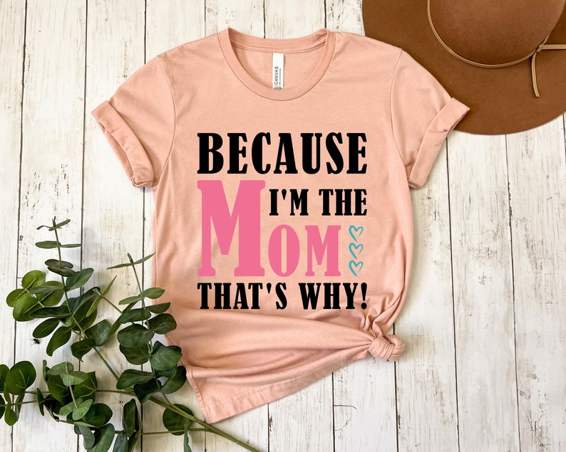 May include: A peach colored t-shirt with the text "Because I'm the Mom That's Why!" in black and pink lettering. The word "Mom" is in pink and has three small pink hearts above it.