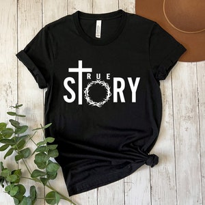 Op de afbeelding: Zwart T-shirt met een wit grafisch ontwerp van een kruis en een doornenkroon met de tekst "TRUE STORY".