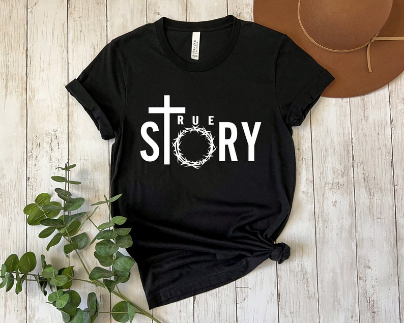 Op de afbeelding: Zwart T-shirt met een wit grafisch ontwerp van een kruis en een doornenkroon met de tekst "TRUE STORY".