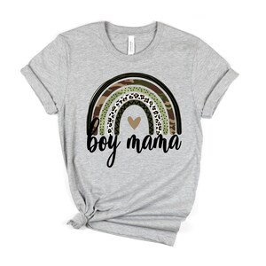 K&ouml;nnte beinhalten: Ein graues T-Shirt mit einem Regenbogen-Design mit Tarnmuster, Leopardenmuster und schwarz-wei&szlig;en Mustern. Der Regenbogen hat ein kleines Herz in der Mitte. Der Text "boy mama" ist unter dem Regenbogen gedruckt.