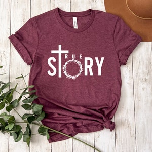 Op de afbeelding: Een maroon t-shirt met een wit kruis en de woorden "TRUE STORY" erop gedrukt. Het kruis is omringd door een witte krans.