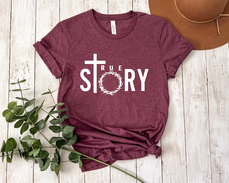 Op de afbeelding: Een maroon t-shirt met een wit kruis en de woorden "TRUE STORY" erop gedrukt. Het kruis is omringd door een witte krans.