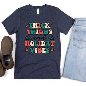 May include: A navy blue t-shirt with the text "THICK THIGHS and HOLIDAY VIBES" in a retro font with red, green, and white accents. The text is surrounded by small Christmas tree and ornament graphics.