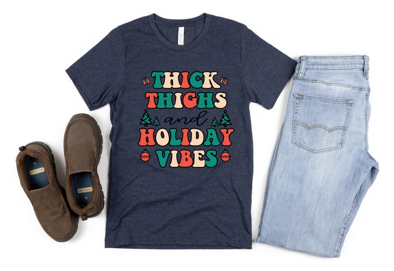 May include: A navy blue t-shirt with the text "THICK THIGHS and HOLIDAY VIBES" in a retro font with red, green, and white accents. The text is surrounded by small Christmas tree and ornament graphics.