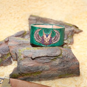 Könnte beinhalten: Grünes Lederarmband mit einem ägyptischen Anubis-Schakal-Kopfdesign. Das Design umfasst Flügel, einen Kragen und einen rot-blauen Kopfschmuck.
