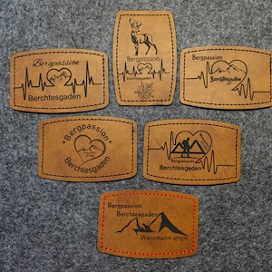 Könnte beinhalten: Sechs braune Lederpatches mit schwarzem Text und Grafiken. Die Patches zeigen den Text "Bergpassion Berchtesgaden" und verschiedene Designs zum Thema Berge, darunter ein Herz mit Bergen, ein Wanderer, ein Hirsch und eine Bergkette.