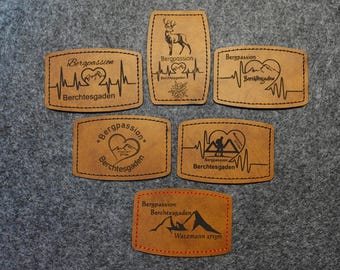 Peut inclure: Six patchs en cuir marron avec du texte noir et des graphiques. Les patchs présentent le texte "Bergpassion Berchtesgaden" et divers motifs sur le thème de la montagne, notamment un cœur avec des montagnes, un randonneur, un cerf et une chaîne de montagnes.