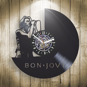 Op de afbeelding: Een zwarte vinyl platenspeler klok met een silhouet van een persoon die in een microfoon zingt. De wijzerplaat van de klok is wit met zwarte cijfers en een gouden uurwerk. De tekst "BON JOVI" is in wit op de plaat gedrukt.