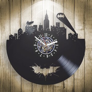 Pode incluir: Um relógio de vinil preto com uma silhueta do logotipo do Batman e uma cidade. O mostrador do relógio é branco com números e ponteiros pretos.
