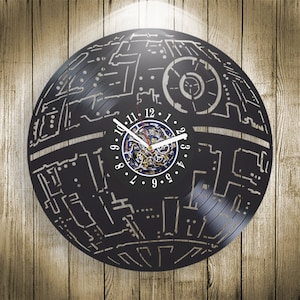 Puede incluir: Un reloj de pared negro hecho de un disco de vinilo, con un diseño detallado que se asemeja a una estación espacial. El reloj tiene manecillas blancas y una esfera central con engranajes visibles. El reloj está montado sobre un fondo de madera.