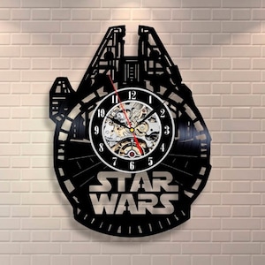 May include: A black vinyl record clock shaped like the Millennium Falcon from Star Wars. The clock face features a silver background with black numbers and hands. The clock is mounted on a white brick wall.