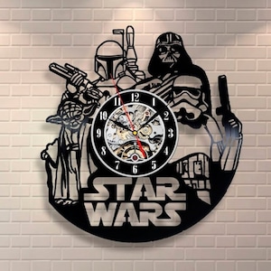 Puede incluir: Reloj de vinilo negro con personajes de Star Wars, incluyendo Darth Vader, Yoda, Boba Fett y un Stormtrooper. La esfera del reloj es un mecanismo de engranaje plateado con números y manecillas negros. El texto "STAR WARS" está impreso en blanco debajo de la esfera del reloj.