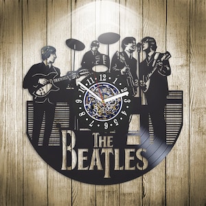 Pode incluir: Relógio de vinil preto e branco com uma imagem dos Beatles tocando instrumentos. O mostrador do relógio é de design circular com algarismos romanos e um mecanismo de relógio detalhado.
