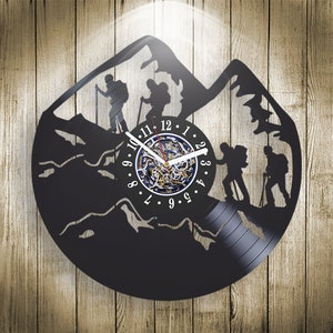 Pode incluir: Relógio de vinil preto com uma cena de montanha e caminhantes. O mostrador do relógio tem um design azul e dourado com algarismos romanos.