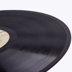 May include: Close-up of a black vinyl record with visible grooves. The record is slightly tilted, showing the edge of the label.