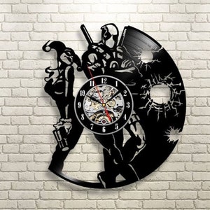 May include: Black vinyl record wall clock featuring a Deadpool and Harley Quinn design. The clock has a visible gear mechanism and a black clock face with white numerals. The figures are cut out of the record.