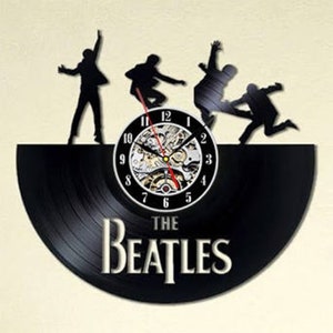 Pode incluir: Um relógio de vinil preto com silhuetas dos Beatles em suas poses icônicas. O mostrador do relógio é branco com números pretos e um mecanismo de relógio prateado. O disco tem o texto "The Beatles" impresso.