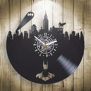 Puede incluir: Reloj de vinilo negro con una silueta de Batman de pie frente a una ciudad. La esfera del reloj es blanca con números y manecillas negros.