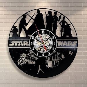 Könnte beinhalten: Eine schwarze Schallplattenuhr mit Figuren aus Star Wars. Das Zifferblatt der Uhr hat einen silbernen Hintergrund mit schwarzen Zahlen und Zeigern. Die Figuren sind Darth Vader, Luke Skywalker, Prinzessin Leia, Han Solo, Chewbacca, R2-D2 und C-3PO. Die Uhr ist von einem schwarzen Hintergrund mit Sternen und den Worten "Star Wars" in silbernen Buchstaben umgeben.