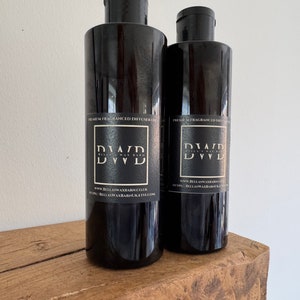 May include: Two black diffuser oil bottles with black caps are displayed on a wooden surface. Each bottle has a label with the text "PREMIUM FRAGRANCED DIFFUSER OIL" and the brand name "DVD". The website address is also visible on the label.