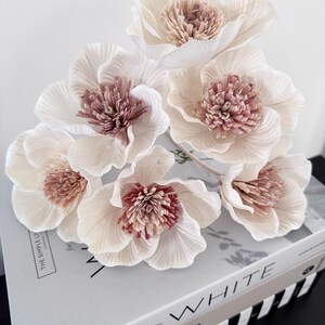 May include: A bunch of artificial flowers with off-white petals and a textured, reddish-pink centre. The flowers are arranged on a stack of books, with the word "WHITE" visible on the spine. The overall aesthetic is soft and elegant.