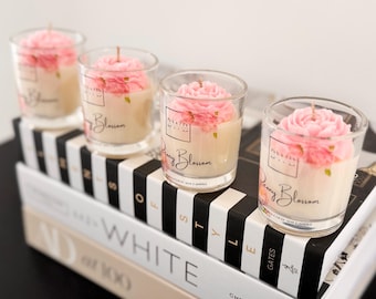 May include: Four clear glass candles with pink flower-shaped wax tops. Each candle features a white label with the text Peony Blossom and a thin wick. The candles are arranged in a row, each containing a cream-colored soy wax base.