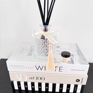 May include: A decorative glass diffuser with black reeds, a white ribbon, and a tassel, sits atop a stack of books. The books are titled "WHITE", "AD at 100", and "THE SIMPLE LIVING". A small black-topped bottle is also present.