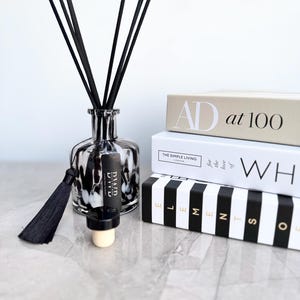 May include: A black and white glass diffuser bottle with black reeds, a black tassel, and a cork. The bottle is next to three books with the titles "AD at 100", "THE SIMPLE LIVING", and "ELEMENTS".