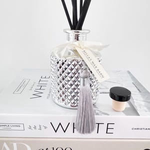May include: A silver diamond-patterned diffuser bottle with black reeds, a silver tassel, and a tag that reads "Bella's Wax Barn". A black-topped cork and a stack of white books complete the display.